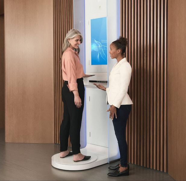 A woman stands on a body scanner smiling at her doctor. The scanner screen shows her breaths-per-minute.